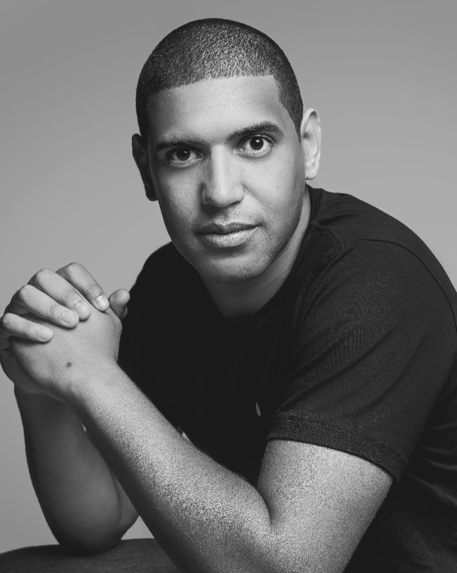 Black and white portrait of a man with his hands clasped, sitting on a textured surface.