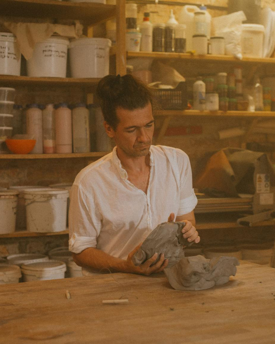 Man working with clay in a pottery studio filled with shelves of pottery and supplies.