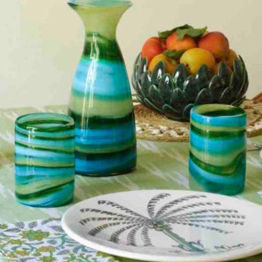 Decorative table setting with green and blue striped glasses, a vase, and a bowl of fruit on a green tablecloth.
