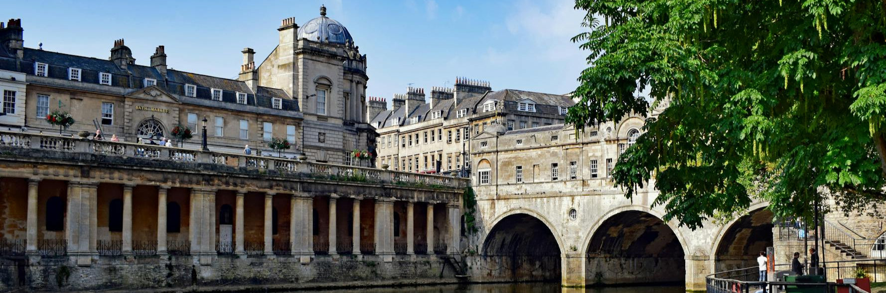 Historic buildings reflected in a calm water body with greenery on the right.