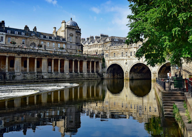 Historic buildings reflected in a calm water body with greenery on a clear day.