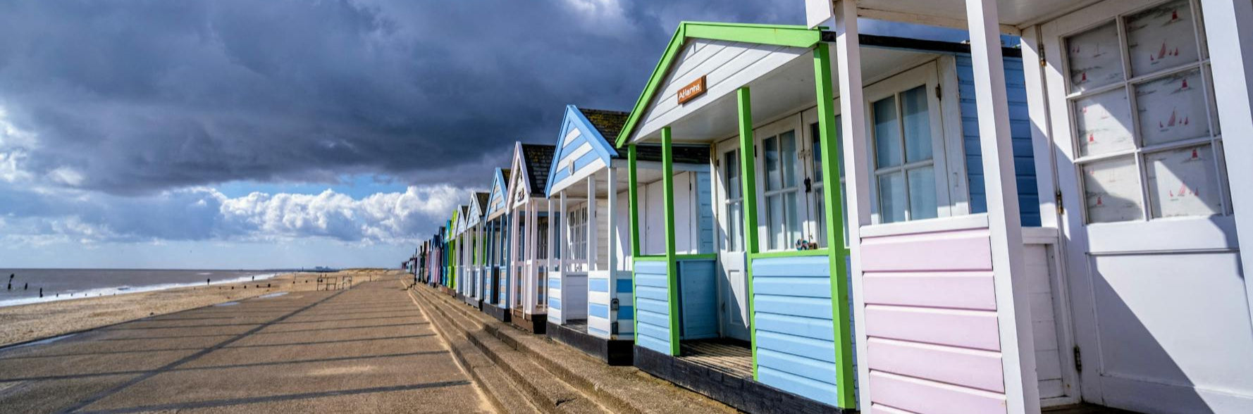 Colorful beach huts along a sandy promenade with a cloudy sky.
